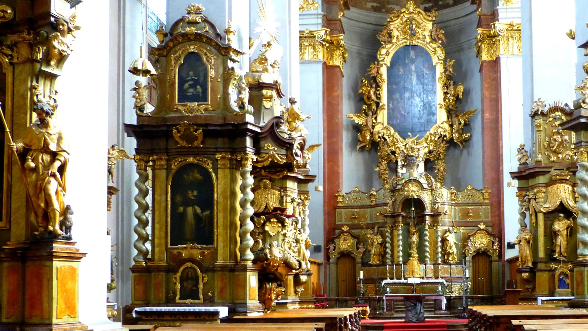 The nave of St Giles' Church in Prague looking towards a gilded high altar framed in gold, with a side altar of twisted columns and carved saints on the left and pale walls above.
